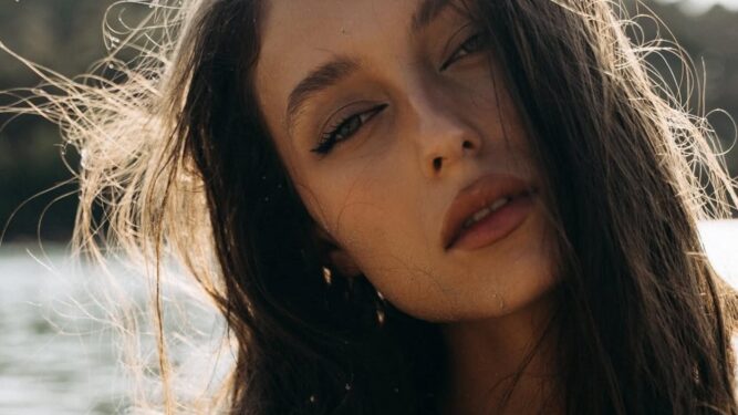 Κοντινό πορτρέτο γυναίκας με βρεγμένα μαλλιά και φυσικό μακιγιάζ, με βλέμμα στραμμένο ελαφρώς προς τα πλάγια, σε εξωτερικό φωτισμό δίπλα σε νερό