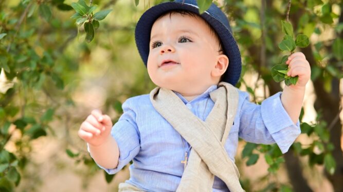 Μικρό παιδί με γαλάζιο πουκάμισο, υφασμάτινο φουλάρι και μπλε καπέλο κάθεται ανάμεσα σε κλαδιά δέντρου, χαμογελώντας και κοιτάζοντας προς τα πάνω